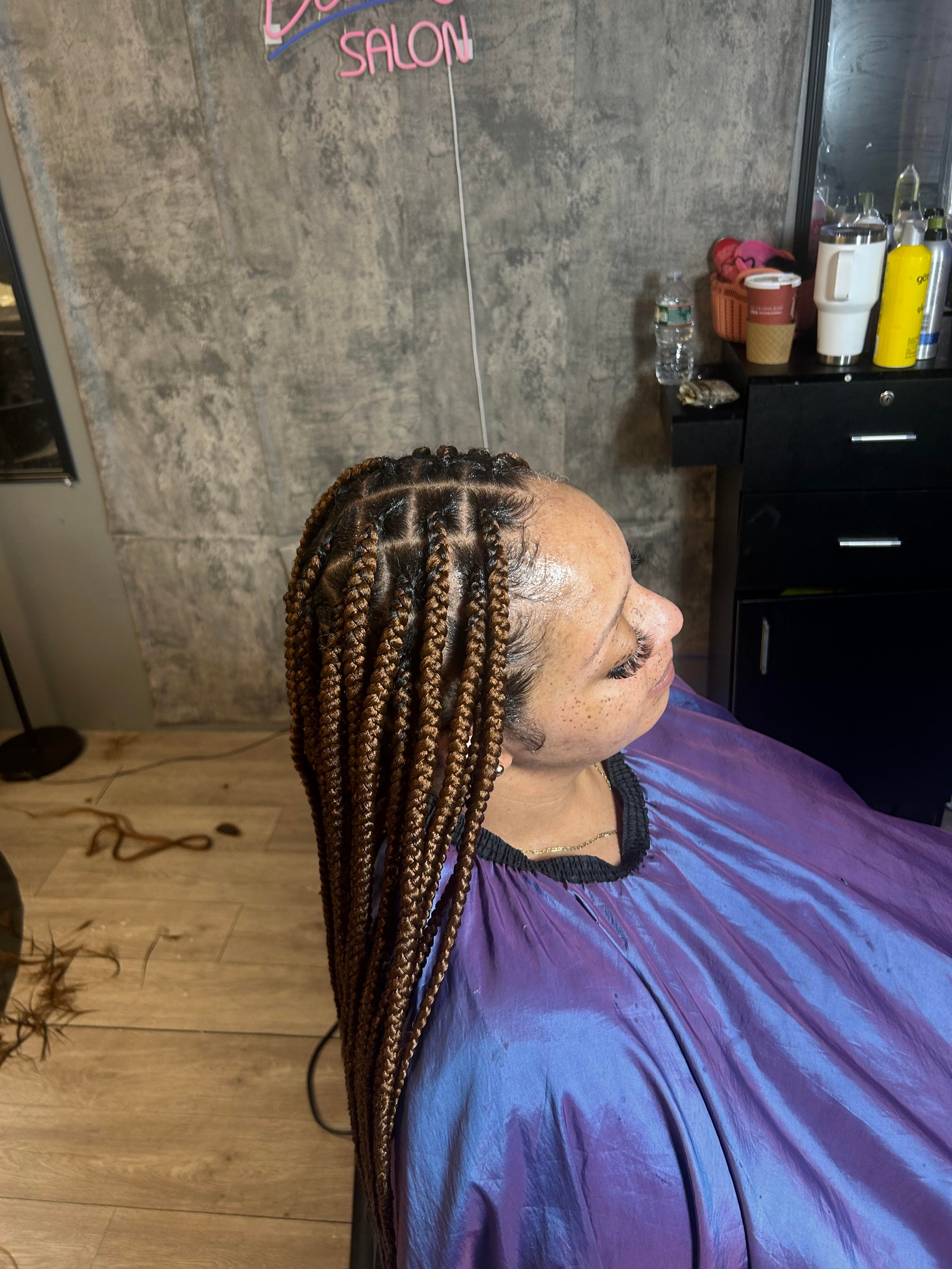 Person with long braided hair sitting in salon chair under neon sign
