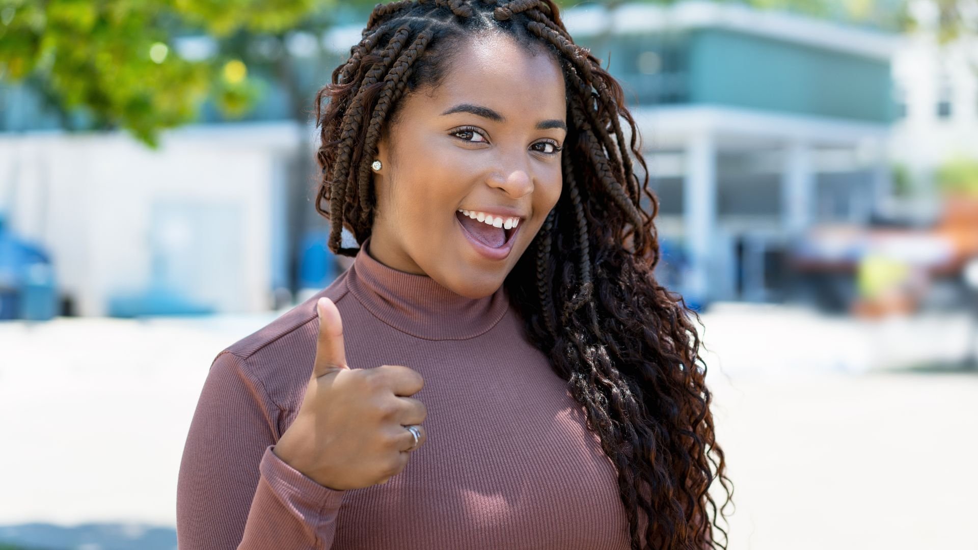 Excited person with long braids giving thumbs up and bright smile outdoors